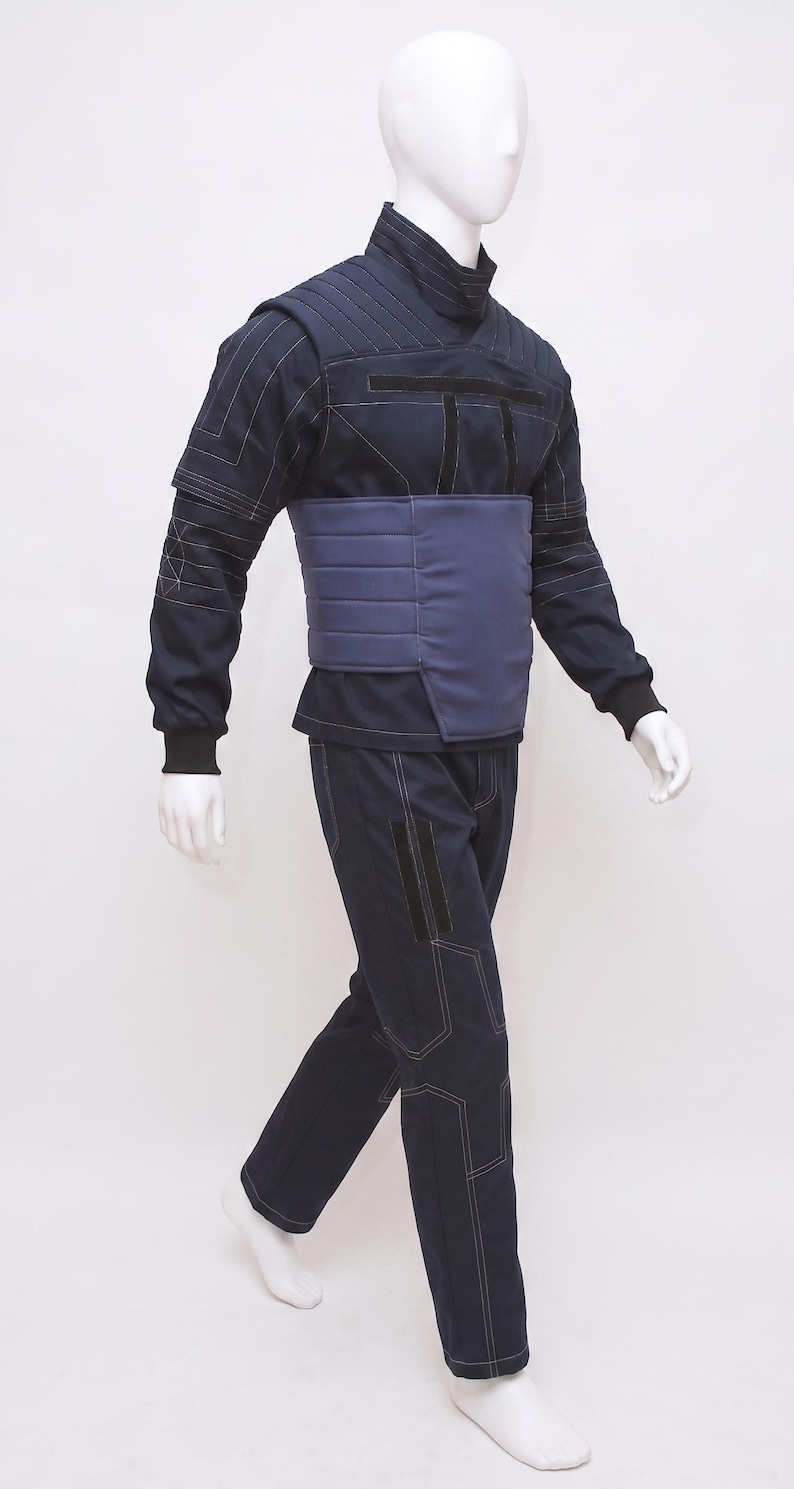 May include: A navy blue jumpsuit on a mannequin. The outfit has a high collar, long sleeves, and a contrasting blue panel across the midsection. White stitching details the seams. The trousers are straight-legged.