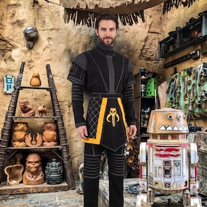 May include: A black long-sleeved tunic and pants set featuring a high collar, white stitched detailing, and a yellow tabard with a black and yellow Mythosaur skull emblem centered on the front panel, worn in a rustic, stone-walled workshop setting.