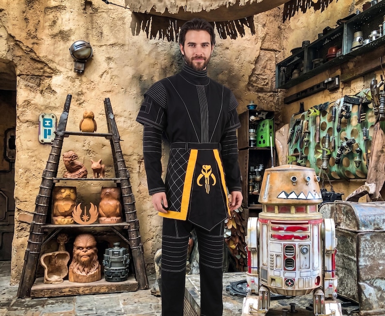 May include: A black long-sleeved tunic and pants set featuring a high collar, white stitched detailing, and a yellow tabard with a black and yellow Mythosaur skull emblem centered on the front panel, worn in a rustic, stone-walled workshop setting.