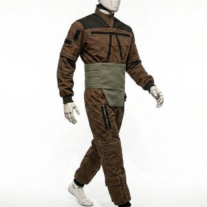 May include: A brown jumpsuit with black accents and a green waist belt is displayed on a mannequin. The outfit features a high collar, quilted detailing, and zippered pockets. The mannequin wears white shoes and has a white head.