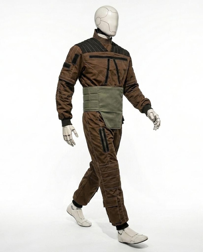 May include: A brown jumpsuit with black accents and a green waist belt is displayed on a mannequin. The outfit features a high collar, quilted detailing, and zippered pockets. The mannequin wears white shoes and has a white head.
