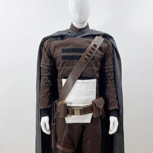 May include: A brown and black costume with a grey cape, displayed on a mannequin. The costume features a leather strap, a white midsection, and a utility belt with pouches. The outfit has intricate stitching details and a high collar.