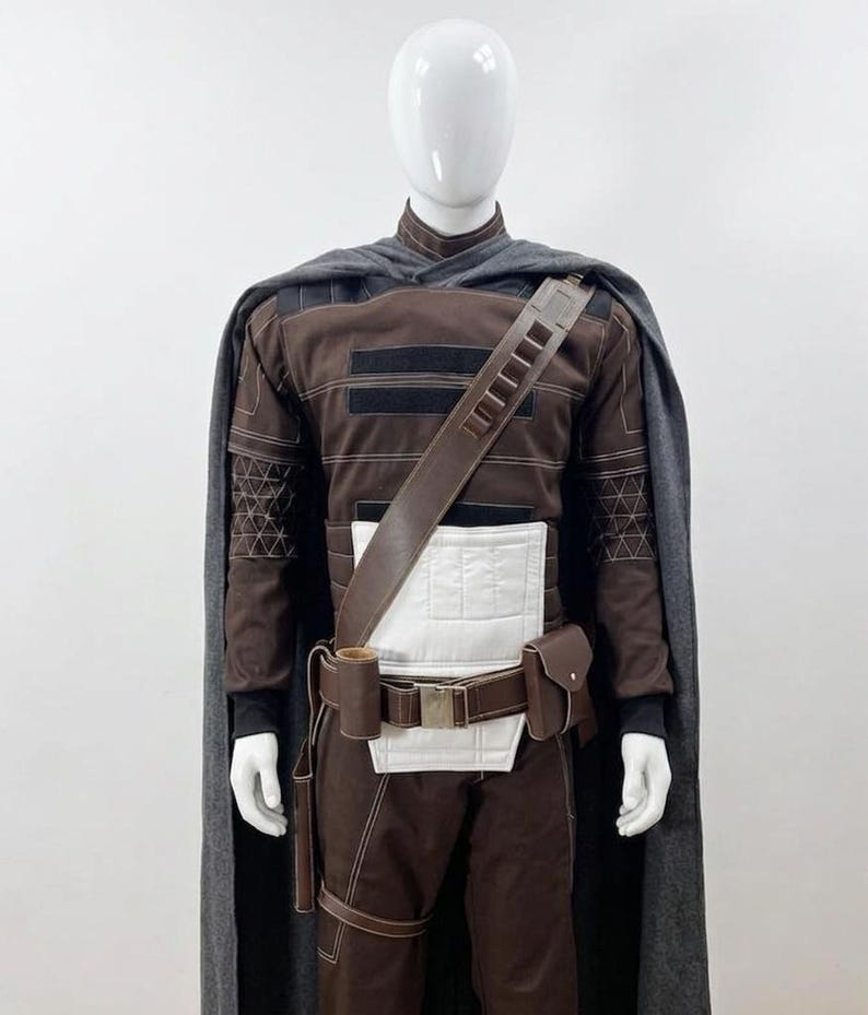 May include: A brown and black costume with a grey cape, displayed on a mannequin. The costume features a leather strap, a white midsection, and a utility belt with pouches. The outfit has intricate stitching details and a high collar.