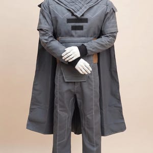 May include: A gray costume displayed on a mannequin. The outfit includes a long coat, pants, and a top with a high collar. White stitching details the seams. The costume has a cape and a belt with a brown leather accent.