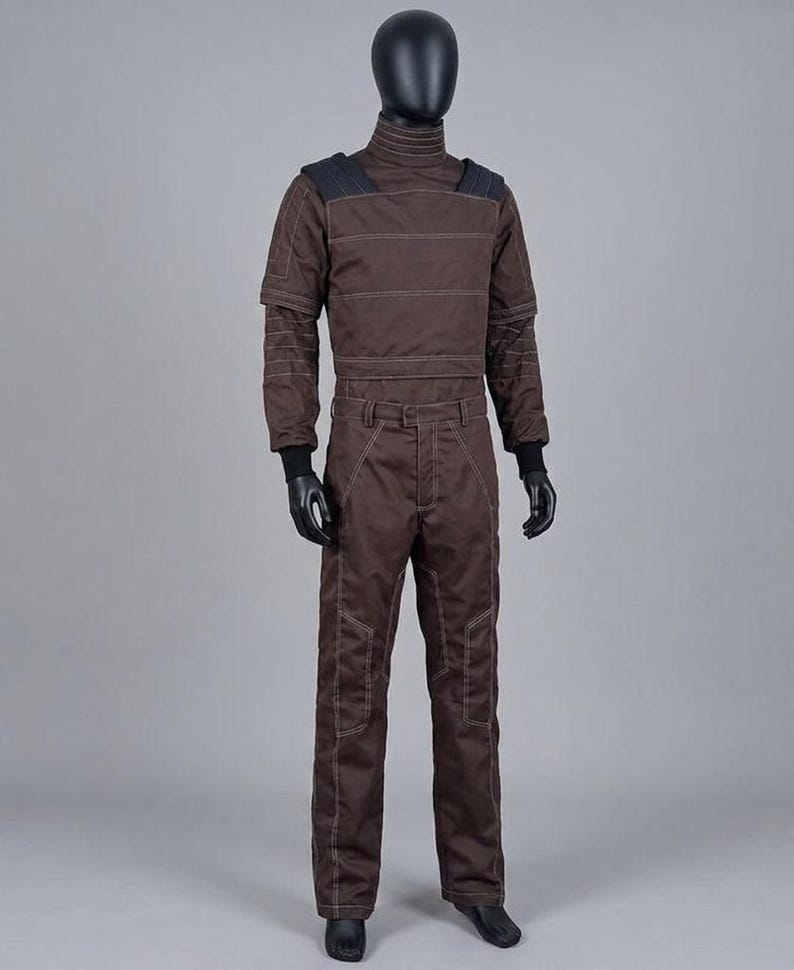 May include: A brown jumpsuit on a mannequin. The outfit has a high collar, reinforced shoulders, and white stitching. The long-sleeved top and trousers are made of a durable material. Black gloves and shoes complete the look.