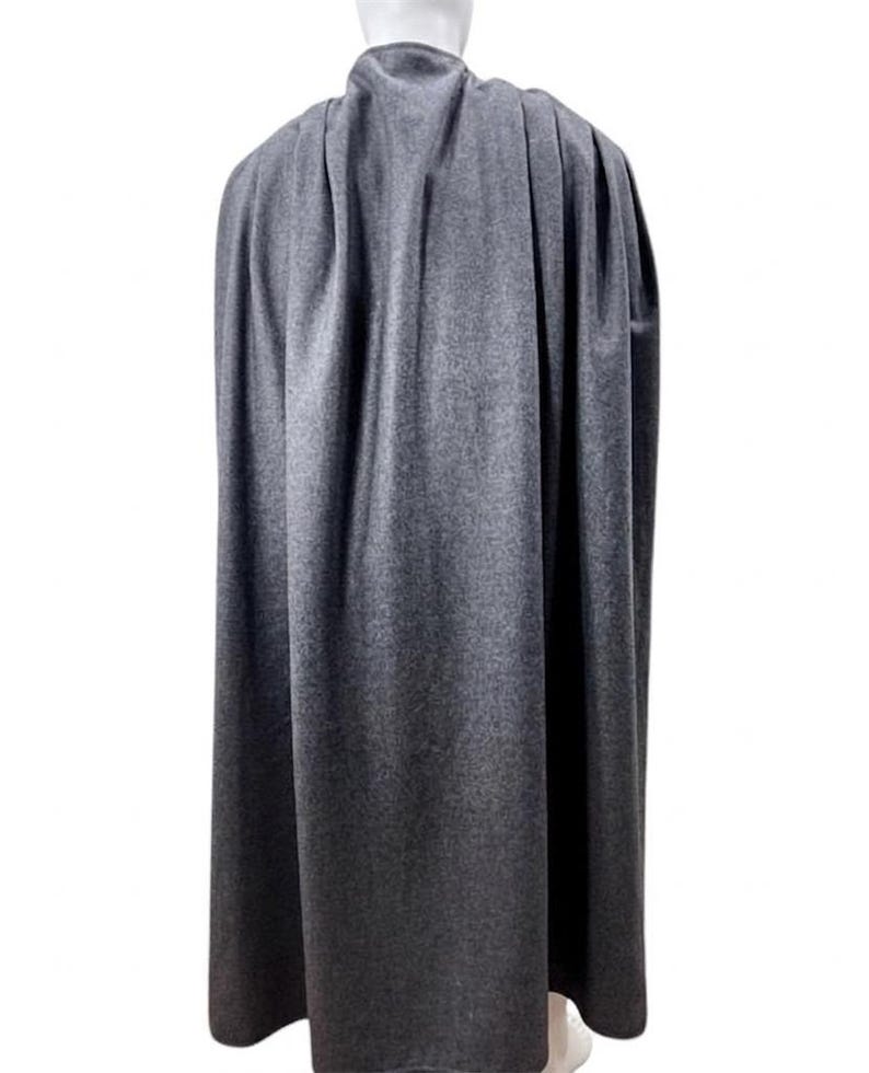 May include: A grey cape made of a soft fabric, draped over a mannequin. The cape is long, reaching to the mannequin's ankles, and has a high collar. The fabric has a subtle texture, and the cape is designed to flow.