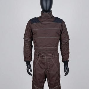 May include: Brown racing suit with white stitching details, displayed on a mannequin. The suit features a high collar, shoulder accents, and long sleeves with black cuffs. The trousers have a matching design with a belt and white stitching.