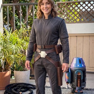 May include: A person in a grey jumpsuit with brown leather accessories, including a belt, pouches, and holsters. A silver buckle is on the belt. A droid with blue and purple accents stands nearby. The setting is outdoors.