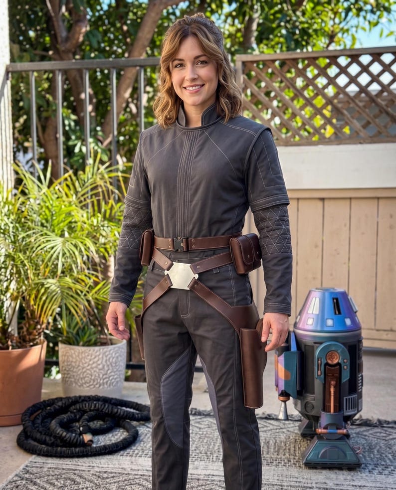 May include: A person in a grey jumpsuit with brown leather accessories, including a belt, pouches, and holsters. A silver buckle is on the belt. A droid with blue and purple accents stands nearby. The setting is outdoors.