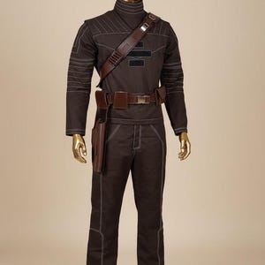May include: A dark brown long-sleeved shirt and matching trousers set featuring white contrast stitching, a high collar, and a brown utility belt with a holster. The outfit includes a brown shoulder strap with metallic accents and black rectangular patches.