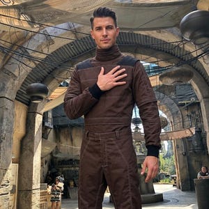May include: A brown jumpsuit with a high collar and black cuffs. The jumpsuit features white stitching and a matching belt. The person is standing in an outdoor setting with a stone archway and hanging lights.