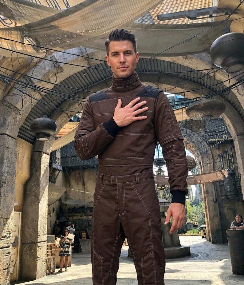 May include: A brown jumpsuit with a high collar and black cuffs. The jumpsuit features white stitching and a matching belt. The person is standing in an outdoor setting with a stone archway and hanging lights.