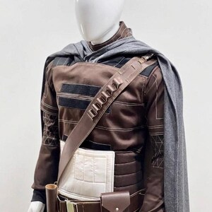 May include: A brown and black costume displayed on a mannequin. The outfit features a long-sleeved top with black detailing, a utility belt with pouches, and a grey cape. The costume has a futuristic or fantasy theme.