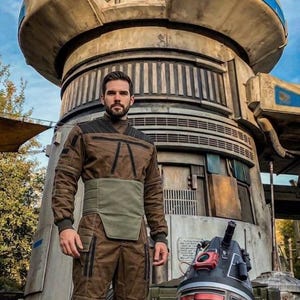 May include: A person in a brown jumpsuit and boots stands next to a droid in front of a large, industrial-looking structure. The droid is primarily black and silver with red accents. The structure is beige and gray with blue accents.