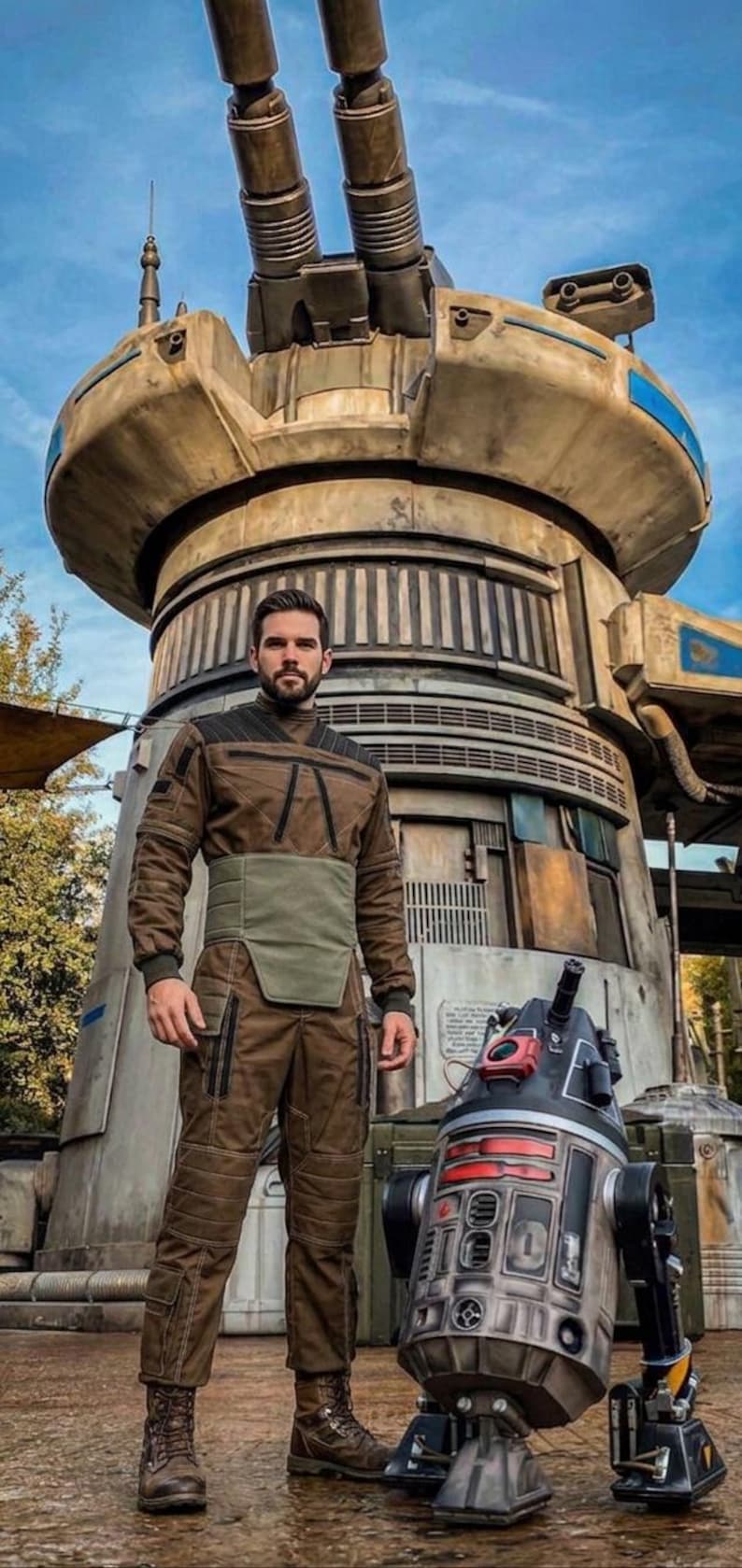 May include: A person in a brown jumpsuit and boots stands next to a droid in front of a large, industrial-looking structure. The droid is primarily black and silver with red accents. The structure is beige and gray with blue accents.