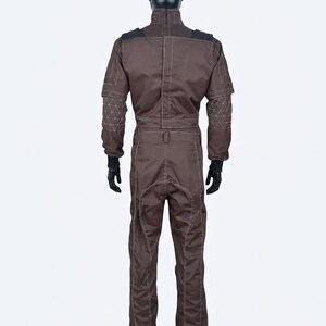 May include: A brown jumpsuit with white stitching details, displayed on a mannequin. The suit features black accents on the shoulders and geometric patterns on the sleeves. The mannequin is wearing black gloves.