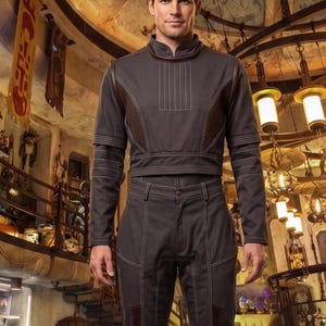 May include: A person in a dark grey outfit with white stitching, comprising a long-sleeved top and trousers. The outfit has a high collar and is worn with brown boots. The background shows a detailed interior with hanging lights.