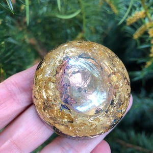 Puede incluir: Un objeto redondo de vidrio transparente con pan de oro incrustado. El objeto tiene una superficie reflectante y se sostiene en una mano sobre un fondo verde borroso. El pan de oro le da al objeto una apariencia brillante y texturizada.