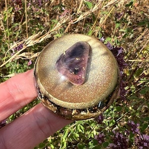 Könnte beinhalten: Ein kreisförmiger Briefbeschwerer aus Kunstharz mit goldener Glitzeroptik und einem polierten, dreieckigen violetten Edelstein in der Mitte. Die Basis enthält dunkle, metallisch wirkende Fragmente, die durch das klare, goldfarbene Harz sichtbar sind.