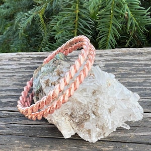 Può includere: Due braccialetti circolari in rame caratterizzati da una texture intrecciata simile a una corda. I braccialetti poggiano su un grande gruppo irregolare di cristalli di quarzo trasparenti e traslucidi con bordi appuntiti e base grezza.