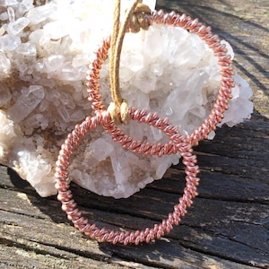 Puede incluir: Dos aros de alambre de cobre, uno más grande que el otro, están trenzados juntos y colocados sobre un lecho de cristales de cuarzo blanco. Los aros están conectados por un cordón de cuero marrón.