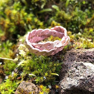 Può includere: Un anello in pelle intrecciata di colore oro rosa. L'anello è appoggiato su un letto di muschio e una roccia.