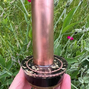 May include: A cylindrical copper tube attached to a circular black base. The base features a textured rope-like border and is embedded with gold-colored flakes. The copper surface has a brushed metallic finish and a vertical orientation.