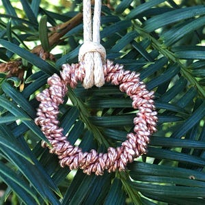 Op de afbeelding: Een koperkleurig, cirkelvormig ornament met een gedraaid touwontwerp, opgehangen aan een koord van natuurlijke vezels. Het ornament hangt tegen een achtergrond van groen gebladerte, wat een natuurlijke, rustieke uitstraling creëert.