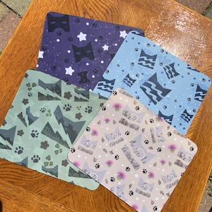 Puede incluir: Un conjunto de cuatro manteles individuales cuadrados con temática de gatos. Los manteles individuales presentan varios patrones, incluyendo siluetas de gatos, huellas de patas, estrellas, montañas y diseños florales. La paleta de colores incluye tonos de azul, verde y beige.