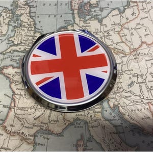 May include: A round, silver compact mirror featuring the Union Jack flag design in red, white, and blue. The mirror is set against a vintage map background, showcasing a detailed cartographic design.