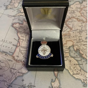 Op de afbeelding: Een HM Armed Forces Veteran-speld in een zwarte presentatiedoos. De speld heeft een wit midden met blauwe en gouden accenten en de woorden "HM Armed Forces Veteran". De doos is open en toont de speld op een zwarte fluwelen inzet.