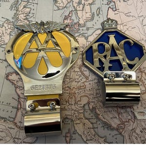 Könnte beinhalten: Zwei Oldtimer-Autoabzeichen, eines mit gelbem und silbernem Design und den Buchstaben "AA", das andere mit blauem und silbernem Design und den Buchstaben "RAC". Beide Abzeichen haben eine silberne Halterung. Der Hintergrund ist eine Vintage-Karte.