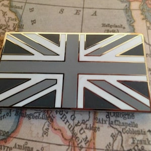 Pode incluir: Um alfinete retangular com um design estilizado da Union Jack em tons de cinza, branco e preto, com uma borda dourada. O alfinete está sobre um mapa vintage.