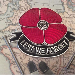 Peut inclure: Un emblème de coquelicot rouge avec l'inscription "LEST WE FORGET" sur une bannière noire. L'emblème est posé sur un fond de carte vintage, avec deux vis argentées de chaque côté.