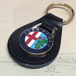 May include: A black leather keyring with a gold-coloured ring. The keyring displays a circular emblem with the words "ALFA ROMEO". The emblem features a red cross, a green snake, and blue and white sections.