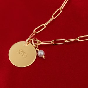 May include: A gold chain necklace with a round pendant featuring the initials "D.S." and a small pearl charm. The necklace is on a red background.