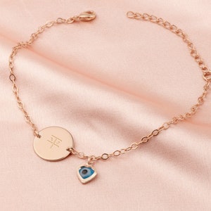 Può includere: Un braccialetto a catena in oro rosa con un ciondolo rotondo inciso con il carattere cinese per "pace" e un ciondolo blu di occhio turco.