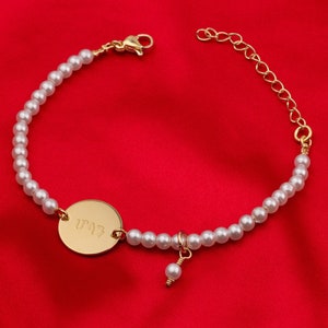 Può includere: Un braccialetto di perle dorate con un ciondolo rotondo in oro che dice "UPAZ" su uno sfondo rosso.