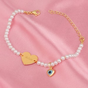 Può includere: Un braccialetto d'oro con un ciondolo a forma di cuore e una catena di perle. Il ciondolo a forma di cuore è inciso con il nome "Emma" ed è accompagnato da un ciondolo a forma di occhio blu.