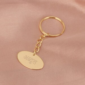 May include: A gold-coloured keychain with a round ring and a chain attached to an oval-shaped tag. The tag has the word "अच्छा" engraved on it.