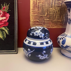 May include: A small, round, dark blue ceramic jar with a lid, decorated with white floral patterns. The jar has a white band around the middle and is placed next to a Delftware vase and a framed needlepoint of a rose.