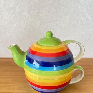 Puede incluir: Un juego de té de cerámica que consiste en una tetera redonda apilada sobre una taza a juego. El conjunto está decorado con rayas horizontales en colores vibrantes del arcoíris, incluyendo rojo, naranja, amarillo, verde, azul y morado, con una tapa verde.