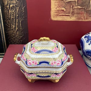 May include: A decorative porcelain trinket box with a lid. The box is white with a yellow and gold lattice pattern, pink roses, and blue ribbon accents. It has gold handles and feet.