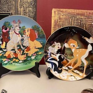 Può includere: Due piatti decorativi con personaggi Disney. Un piatto raffigura i personaggi de "Lilli e il Vagabondo" con sfondo azzurro chiaro. L'altro piatto mostra i personaggi di "Bambi" con sfondo scuro e bordo dorato.