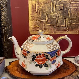 May include: A white porcelain teapot with a floral design featuring orange and red flowers, blue accents, and gold trim. The teapot has a lid with a gold finial and a curved handle. It sits on a wooden slice.