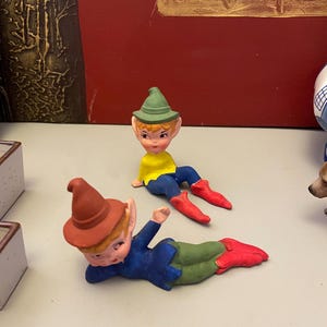 Op de afbeelding: Twee kleine elfenbeeldjes. Eén ligt en de ander zit. Beide hebben een lichte huid, puntoren en dragen hoeden, blauwe shirts, groene broeken en rode schoenen. De achtergrond is een rode muur met gouden accenten.