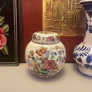 Könnte beinhalten: Ein kleines, rundes, weißes Keramikgefäß mit Deckel, verziert mit einem farbenfrohen Blumenmuster in Rosa-, Orange-, Blau- und Grüntönen. Das Gefäß ist ein dekorativer Gegenstand, möglicherweise zur Aufbewahrung oder Präsentation.