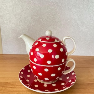Op de afbeelding: Een rode theeset voor één persoon bestaande uit een theepot gestapeld op een theekopje en schoteltje, alles versierd met een wit stippenpatroon. De theepot heeft een witte tuit en handvat, terwijl het theekopje een bijpassend wit handvat heeft.