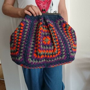 K&ouml;nnte beinhalten: Eine gro&szlig;e, handgefertigte H&auml;keltasche im Granny-Square-Muster. Die Tasche hat einen dunkelblauen Rahmen und ein farbenfrohes Design mit orangefarbenen, lila und grauen Quadraten. Die Tasche wird von einer Person gehalten, die ein wei&szlig;es T-Shirt mit einem Hunde-Motiv tr&auml;gt.