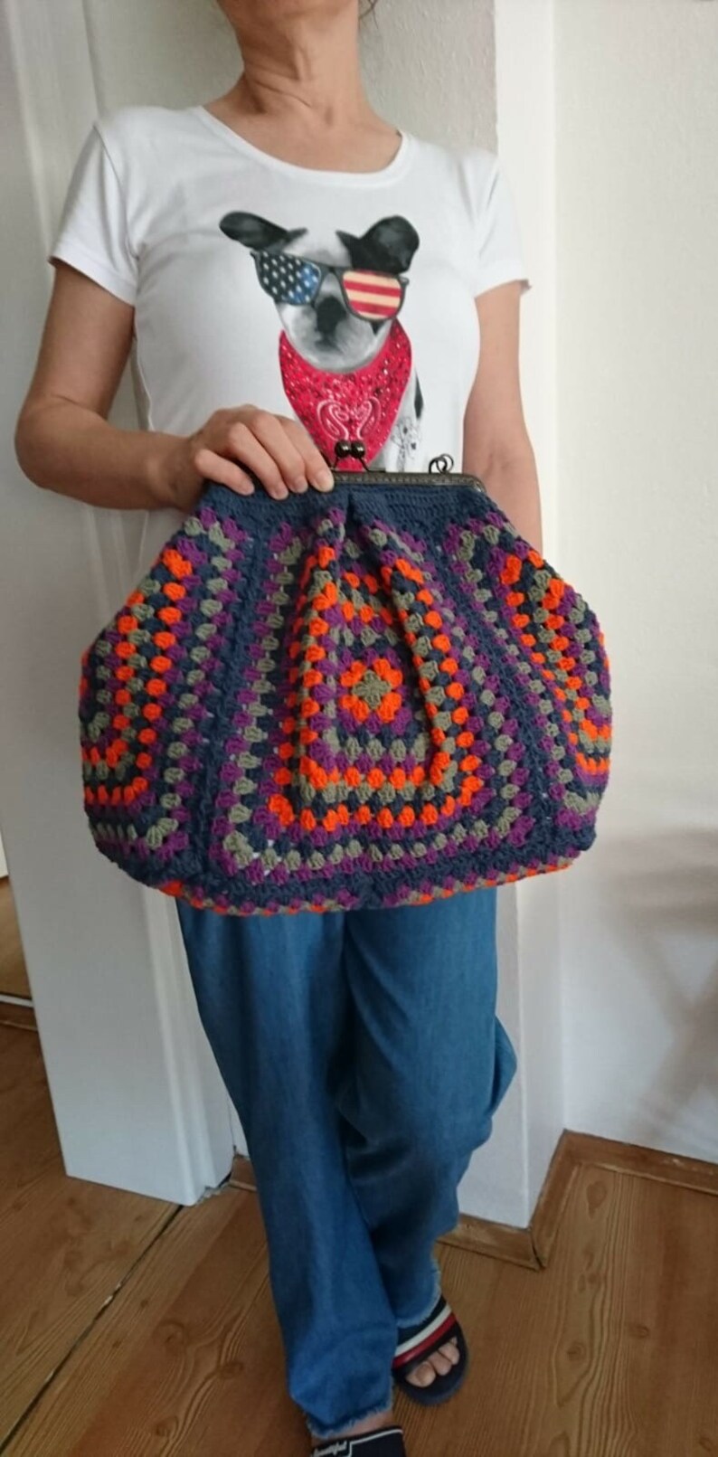 K&ouml;nnte beinhalten: Eine gro&szlig;e, handgefertigte H&auml;keltasche im Granny-Square-Muster. Die Tasche hat einen dunkelblauen Rahmen und ein farbenfrohes Design mit orangefarbenen, lila und grauen Quadraten. Die Tasche wird von einer Person gehalten, die ein wei&szlig;es T-Shirt mit einem Hunde-Motiv tr&auml;gt.